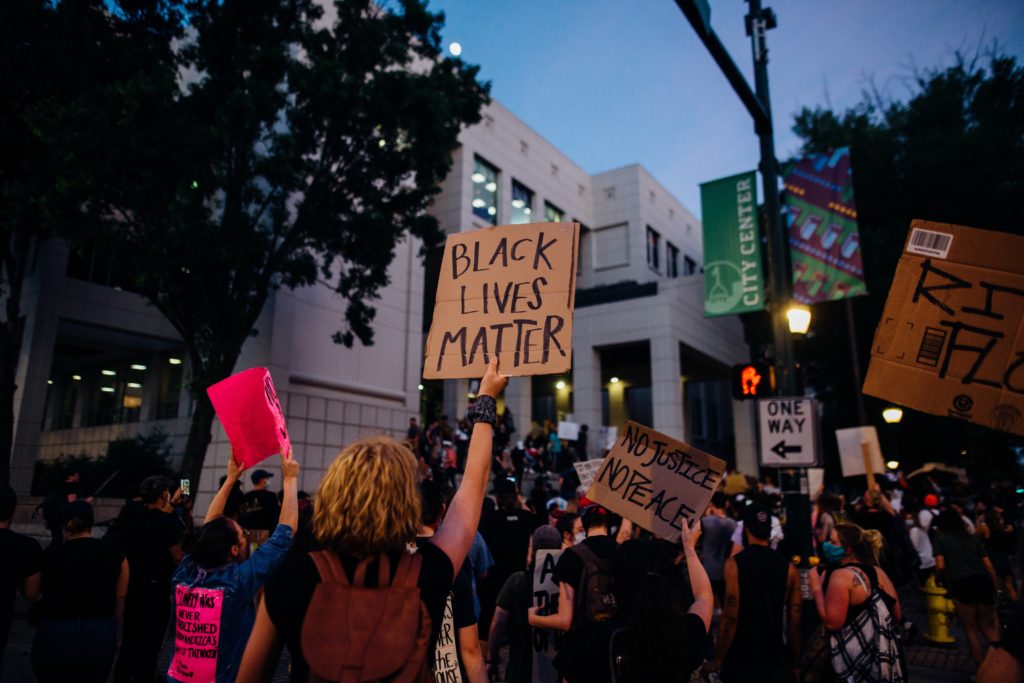 A photo of a crowd of protestors holding up signs. Two cardboard signs can be read, one reads "Black Lives Matter" and the other reads "No Justice No Peace"